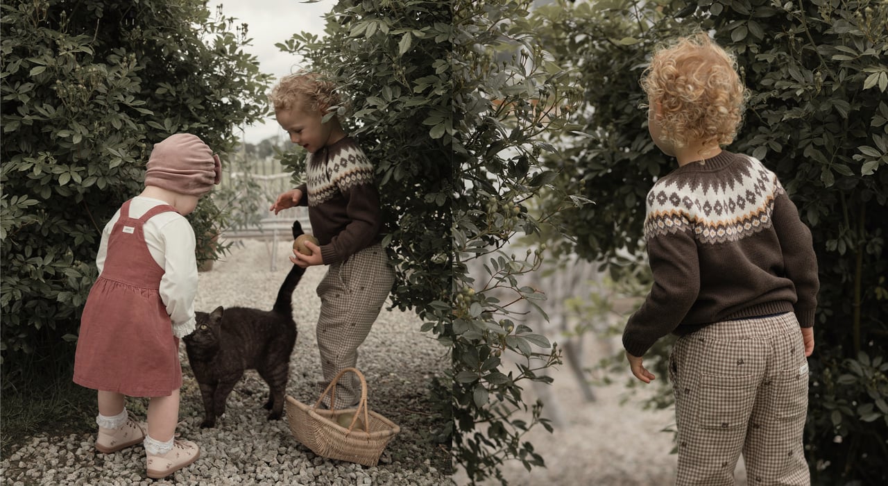 Two children in a pink pinafore dress and a brown patterned knit jumper stand beside a black cat in a garden archway.