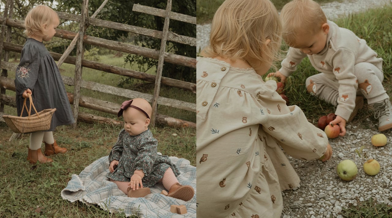 Two young children in patterned dresses outdoors by a rustic wooden fence, with woven baskets and a blanket.