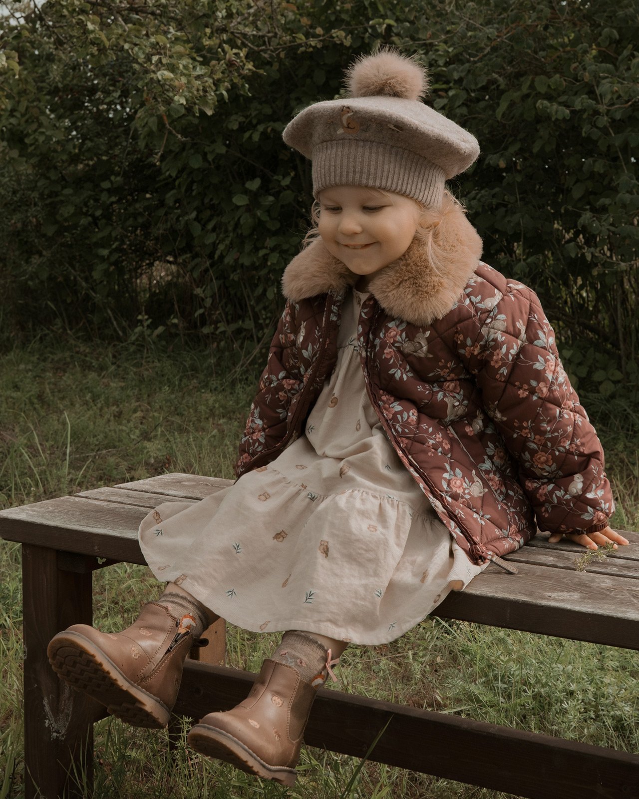 Kind in floraler Steppjacke, Strickbarett, Kleid und braunen Stiefeln im Freien
