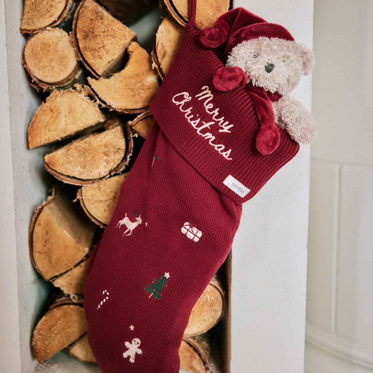 Red Christmas stocking with festive embroidery and a teddy bear, hanging beside stacked firewood