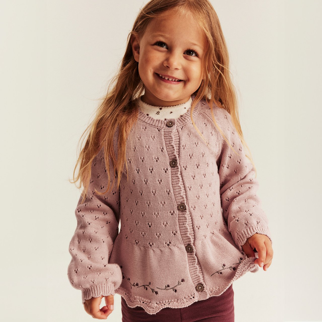 Pink pointelle knit cardigan with buttons, scalloped hem and embroidered floral detail for children.