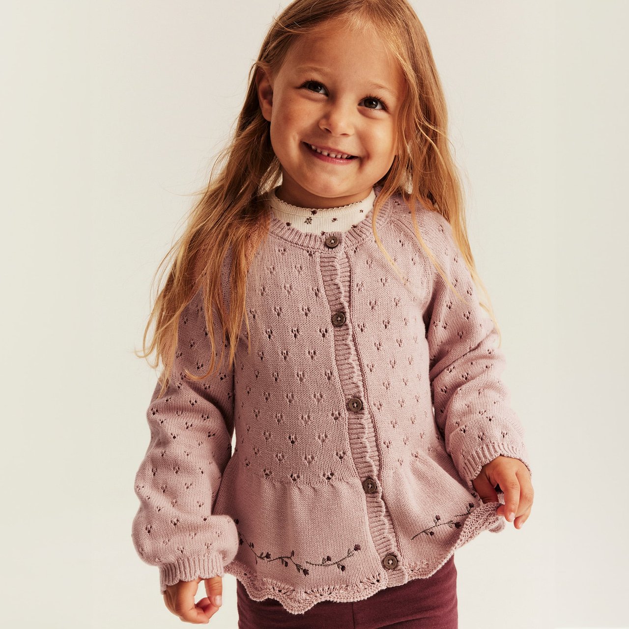Pink pointelle knit cardigan with buttons, scalloped hem and embroidered floral detail for children.