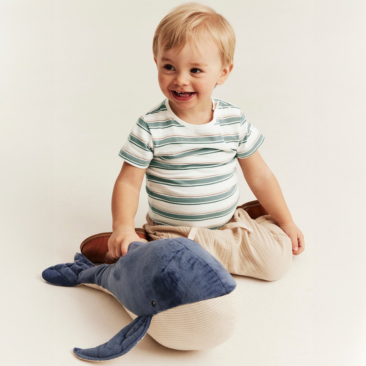 Baby boy wearing a striped short‑sleeve t‑shirt and light trousers, seated on the floor with a soft whale toy.