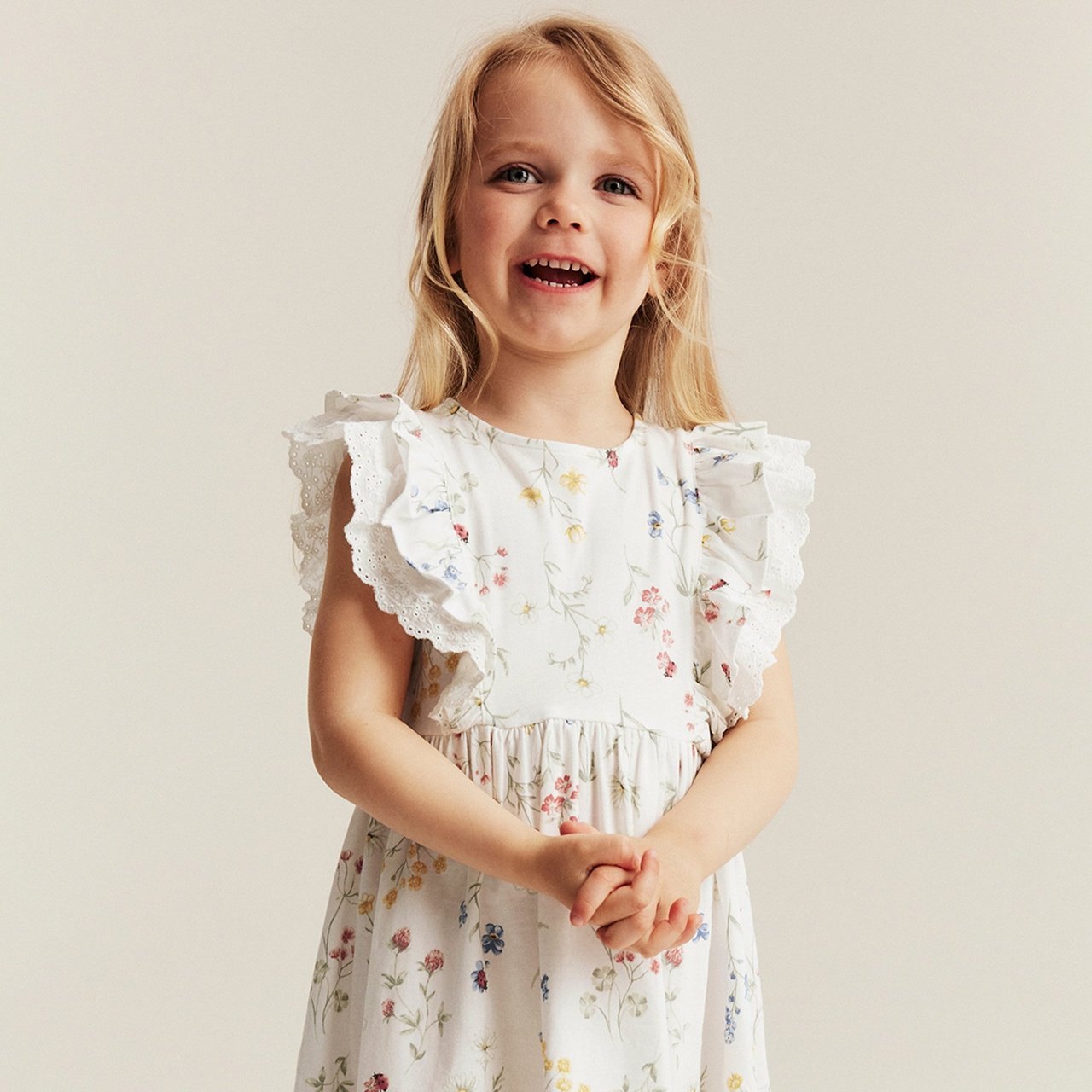 Girl wearing a white sleeveless dress with a soft floral print and ruffle sleeves, photographed against a neutral background.