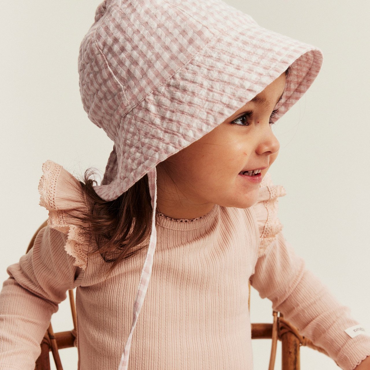 Child wearing a soft ribbed light pink long-sleeve top and a pink gingham hat, seated on a wooden chair.