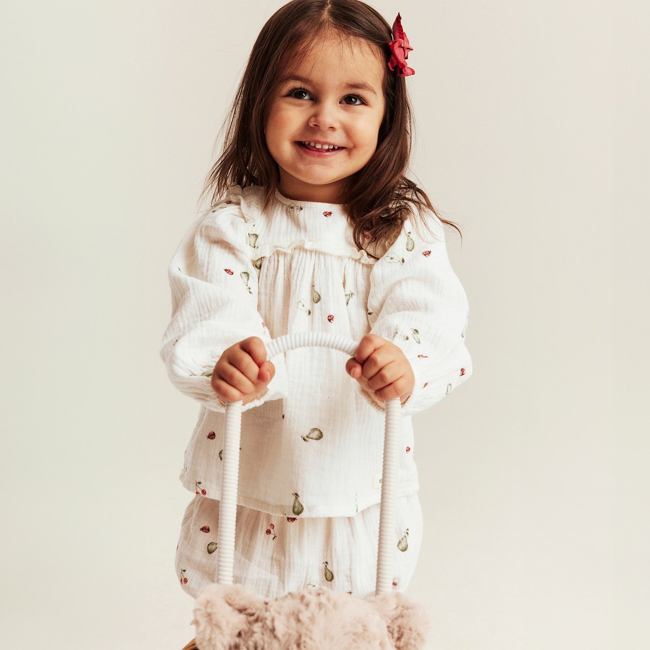 Baby girl wearing a white long-sleeve muslin top and matching trousers with a small print, holding a teddy trolley.