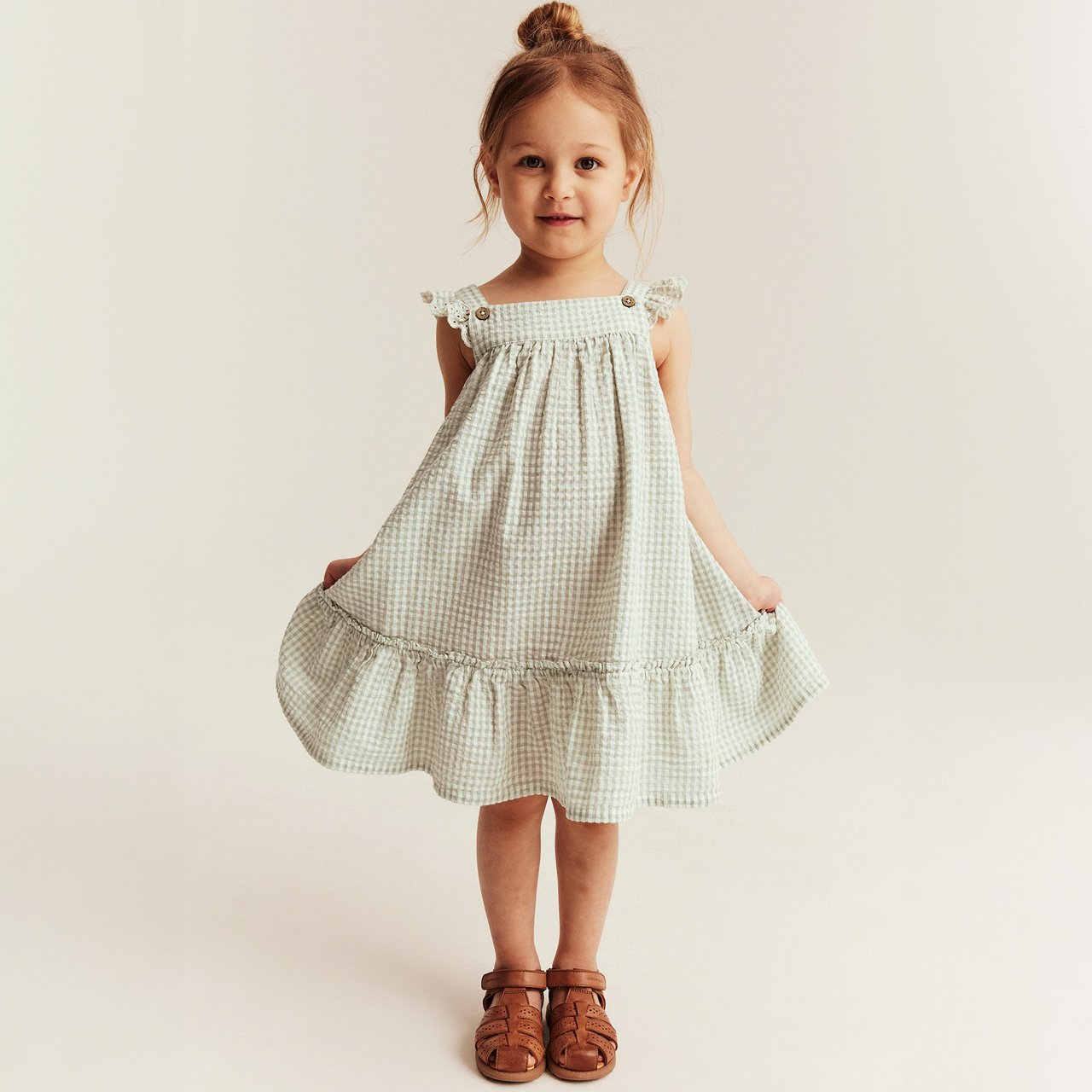 Girl wearing a light green gingham sleeveless dress with ruffle straps, standing against a neutral background.