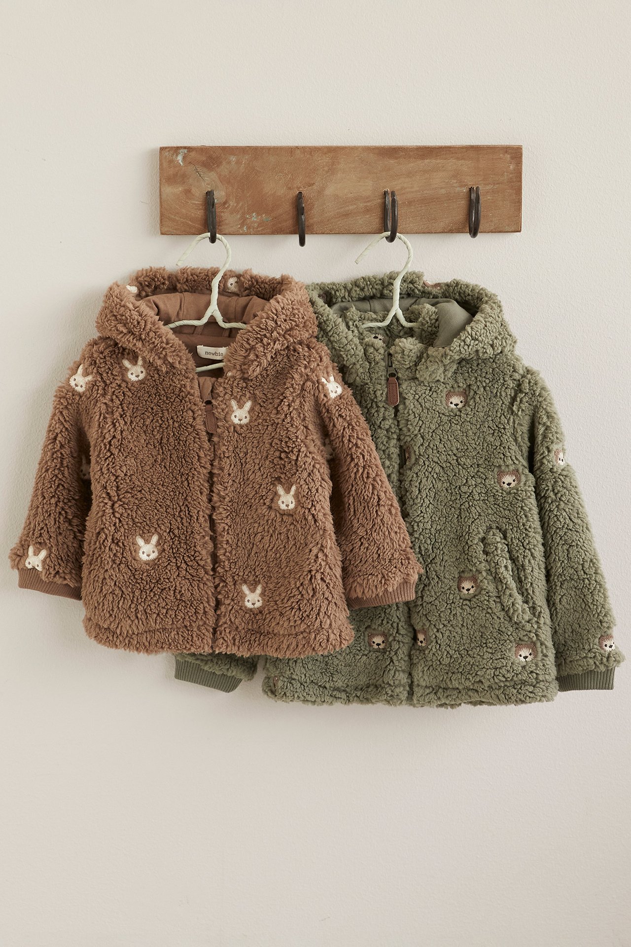 Brown and green teddy fleece jackets with animal motifs hanging on wooden wall hooks.
