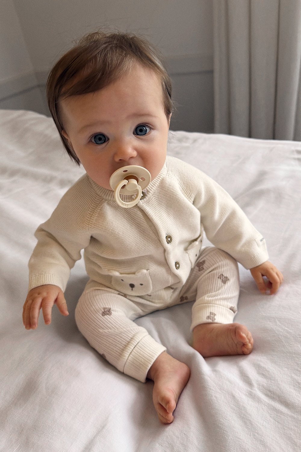 Baby wearing a soft knitted off‑white cardigan and matching trousers with small print, sitting on a light fabric surface.