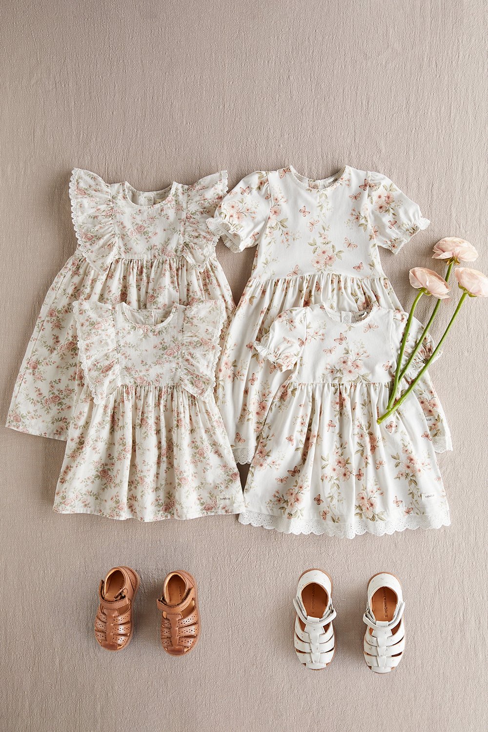 Floral baby and toddler dresses in soft tones laid flat on a neutral background, with small shoes and delicate flowers.