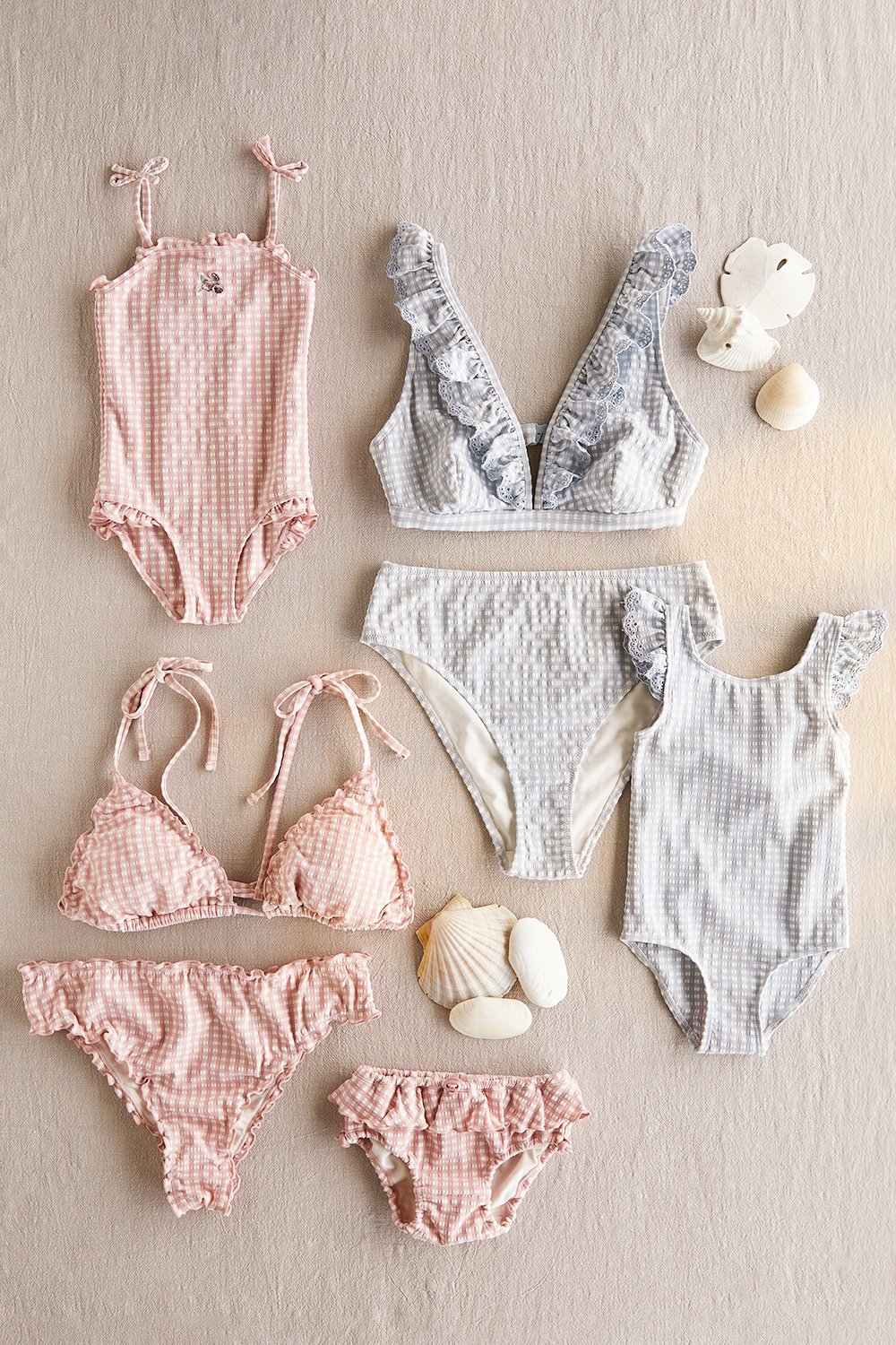 Flat lay of soft seersucker swimwear in light pink and blue, including baby swimsuits and bikinis on a neutral background.