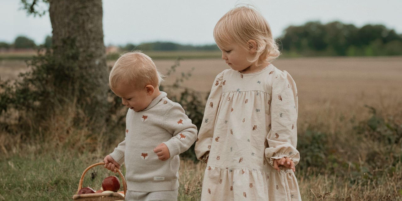 Two young children outdoors, one carrying a basket of apples in a countryside setting