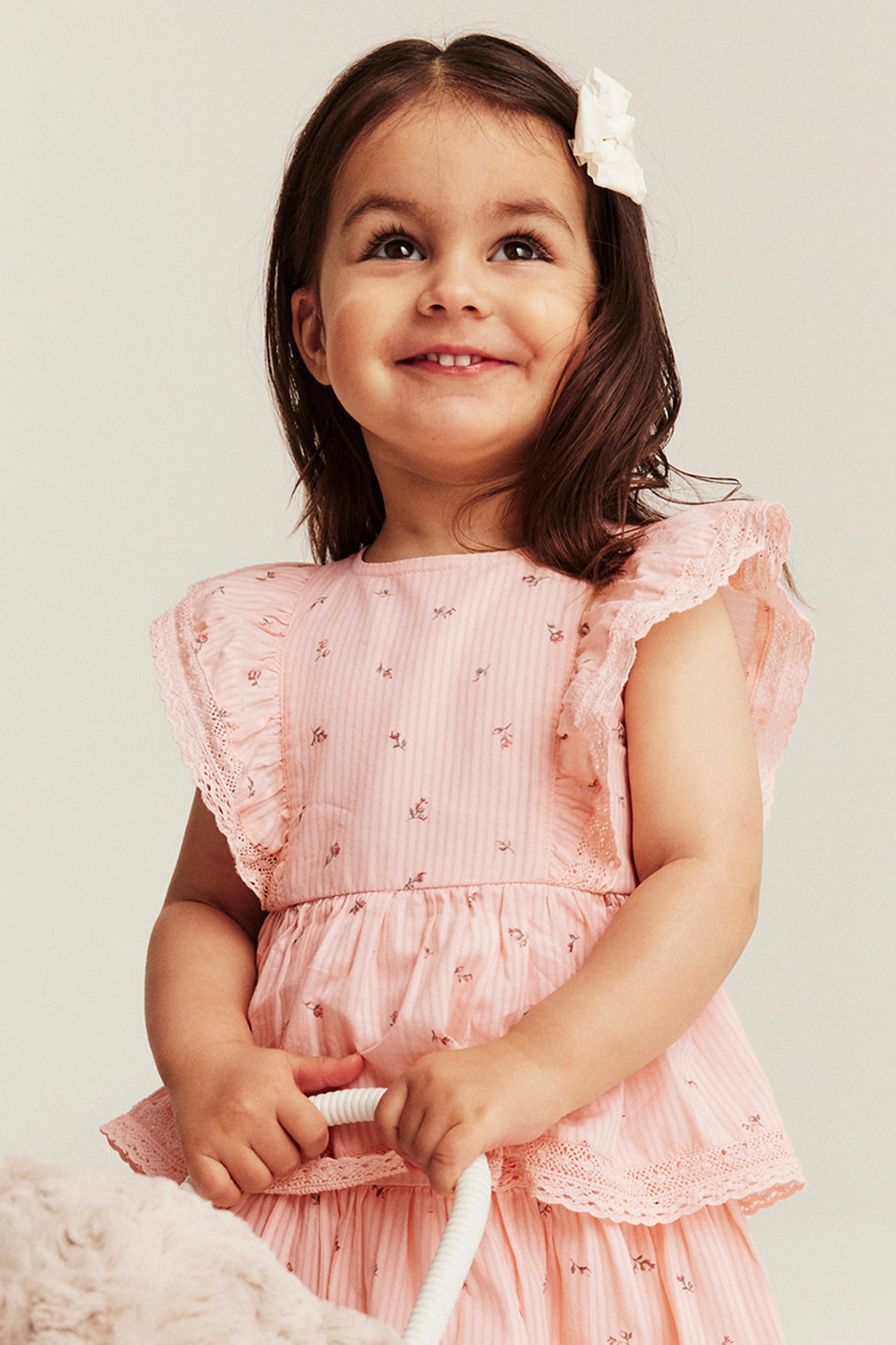 Young child wearing a light pink sleeveless dress with lace frill sleeves, delicate floral print and a white hair bow.