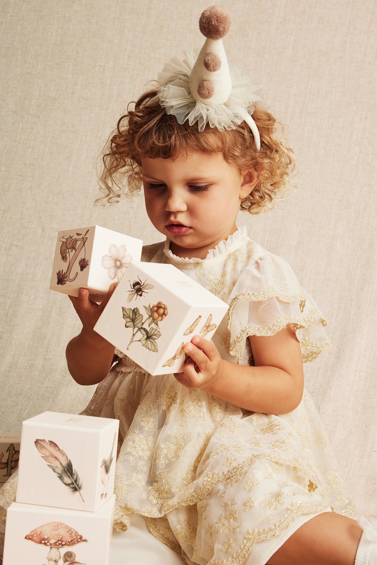 Vorderansicht eines Babys, das cremefarbene gemusterte Papier-Stapelklötze hält, ein cremefarbenes Kleid und einen Partyhut trägt.