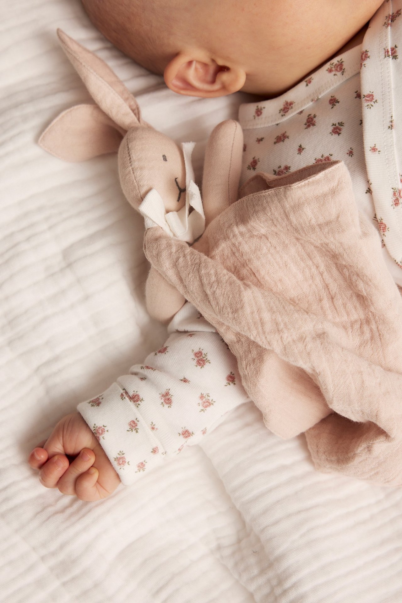 Nahaufnahme eines Babys, das ein hellbeiges Hasen-Schmusetuch hält und ein weißes Langarmshirt mit Blumenmuster trägt.