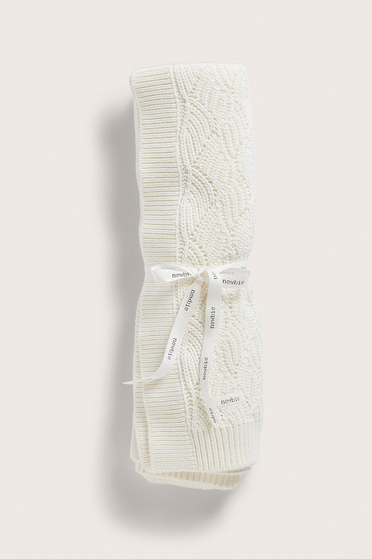 Front view of an off-white knitted baby blanket with a jacquard pattern, tied with a 'Newbie' ribbon.