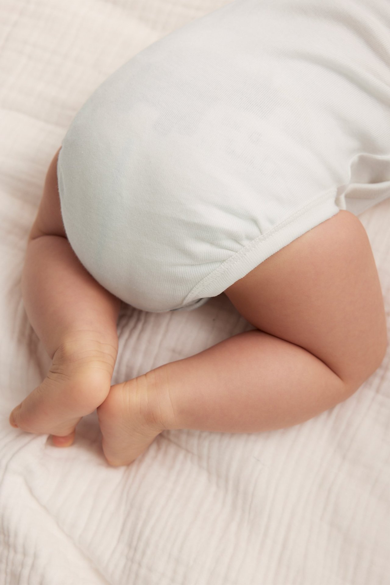 Rückenansicht eines Babys in einem cremefarbenen Baumwoll-Wickelbody, auf einer hellen Decke liegend.
