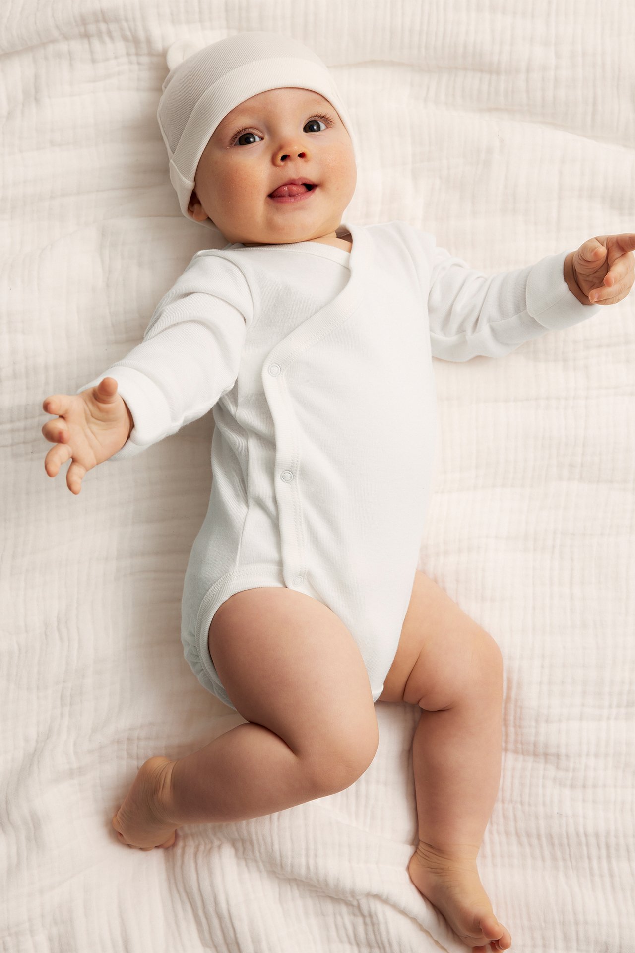 Vorderansicht eines Babys in einem cremeweißen langärmeligen Baumwoll-Wickelbody mit Druckknöpfen und passender Mütze.