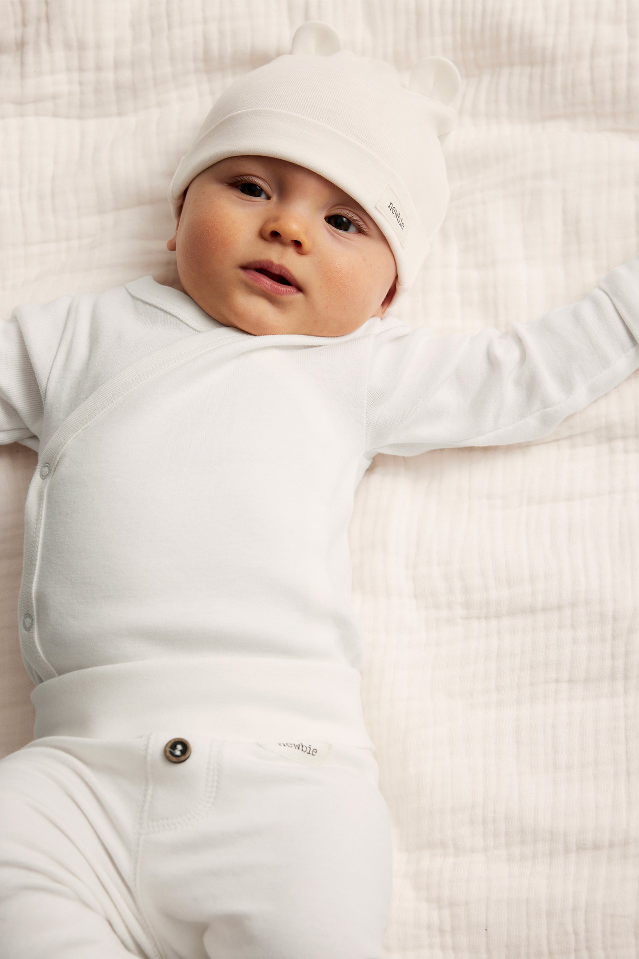 Draufsicht: Baby trägt einen cremeweißen Baumwoll-Wickelbody, passende Mütze mit Ohren und Hose.