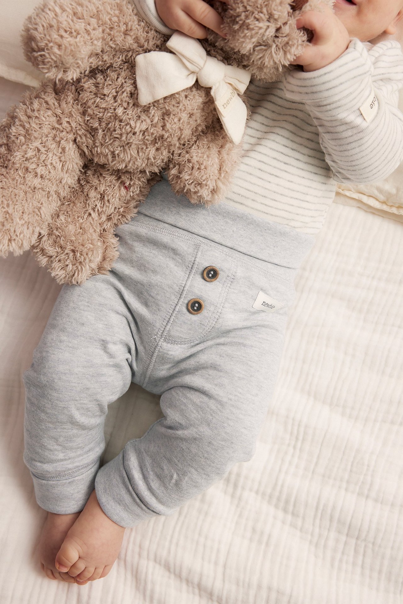 Top view of baby wearing grey melange cotton leggings with decorative buttons, a striped top, and holding a teddy bear.