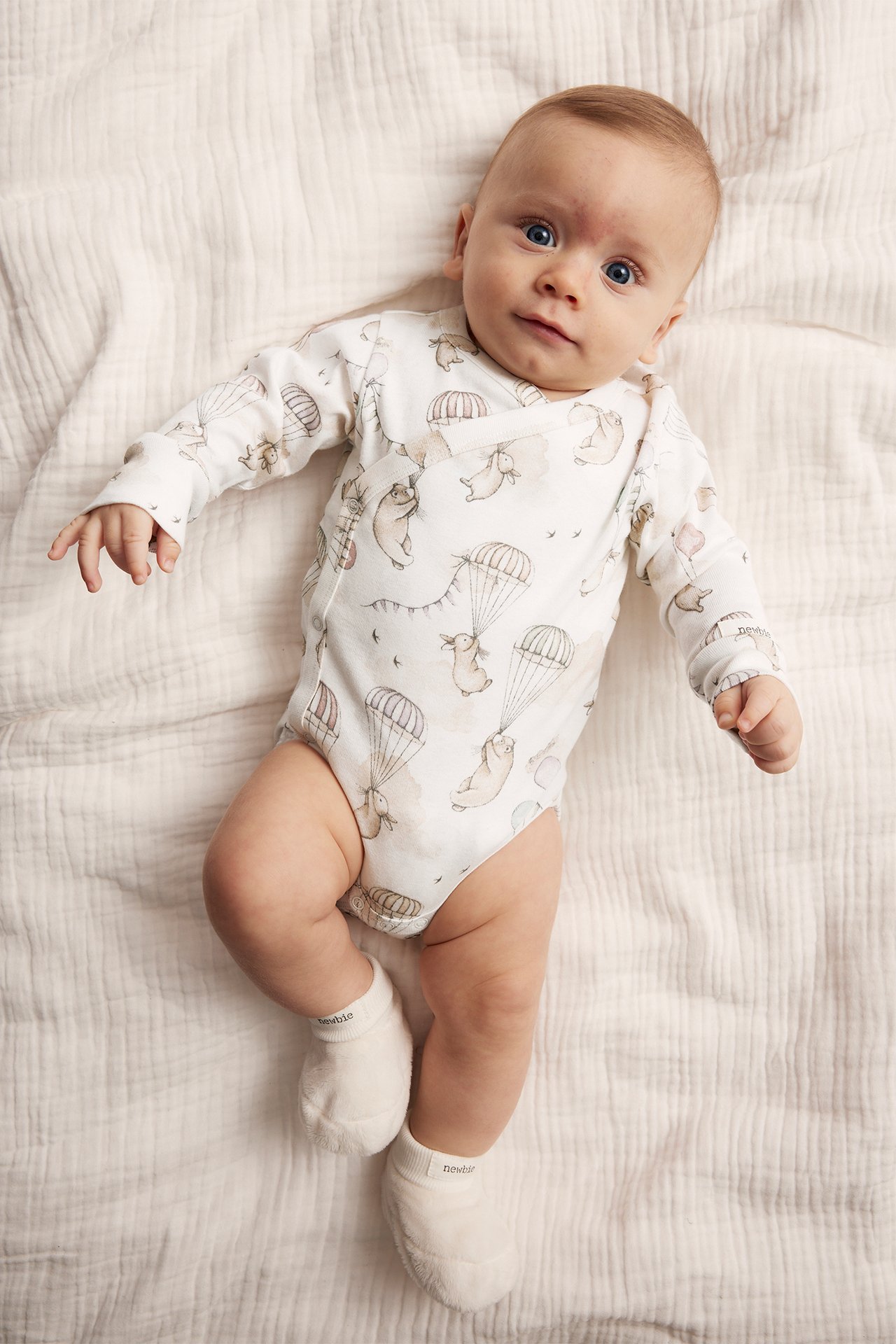 Top-down view of a baby in an offwhite patterned wrap bodysuit with animal motifs and offwhite socks.