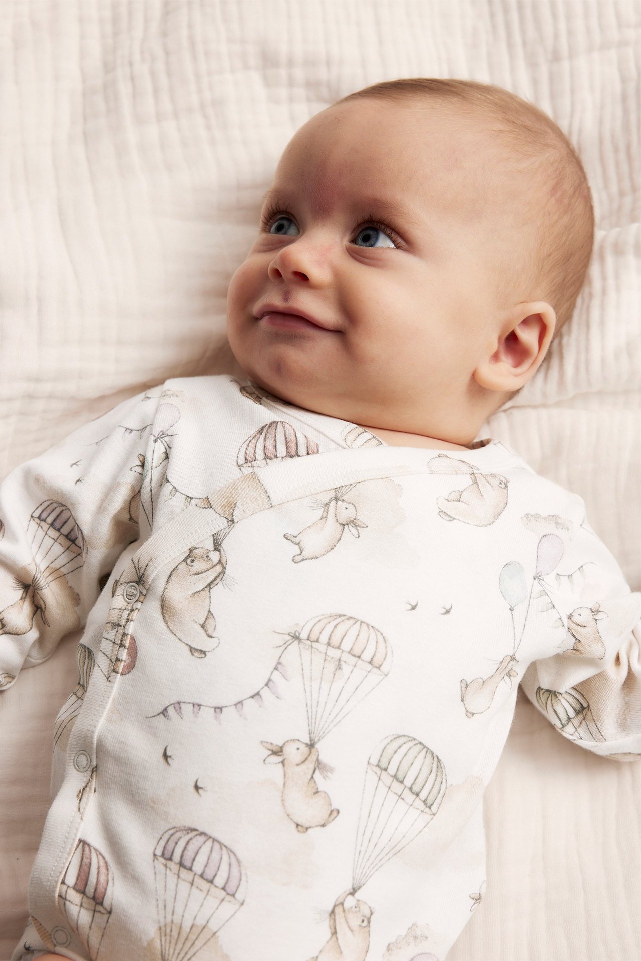 Close-up front view of a baby wearing an off-white patterned wrap bodysuit with rabbit and bear motifs.