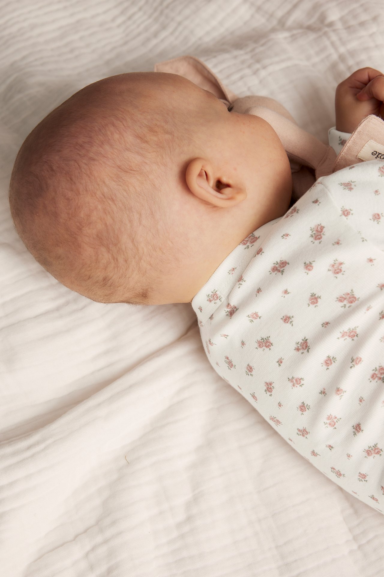Close-up side view of a baby wearing an off-white floral long-sleeved wrap-around bodysuit, holding a pink fabric item.