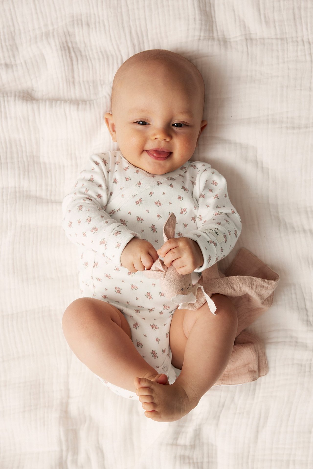 Top-down view of a smiling baby wearing an off-white floral long-sleeved wrap bodysuit, holding a pink bunny comforter.