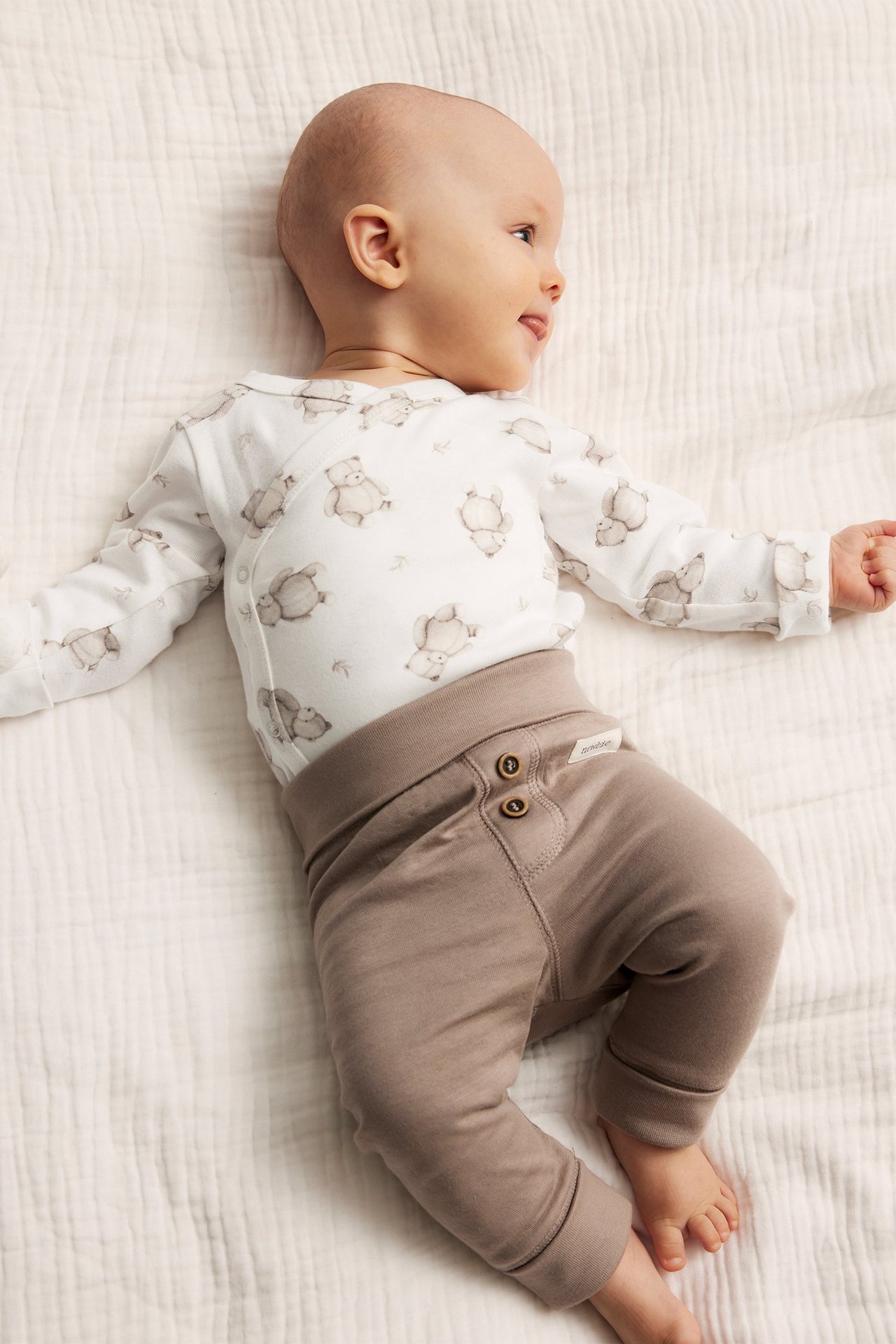 Draufsicht: Baby in off-weißem Wickelbody mit Tiermuster und brauner Hose.