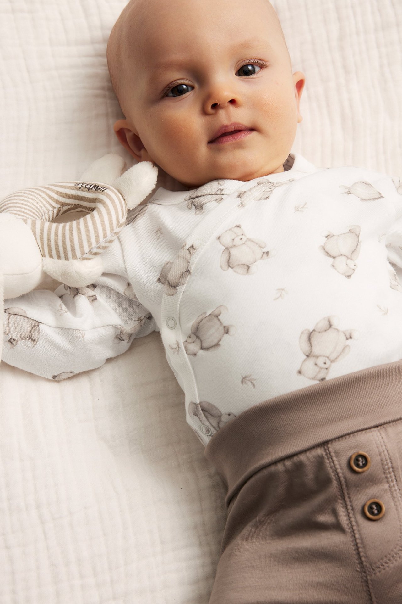 Nahaufnahme eines Babys in cremefarbenem Wickelbody mit Tiermuster, braunen Hosen und gestreiftem Greifling.