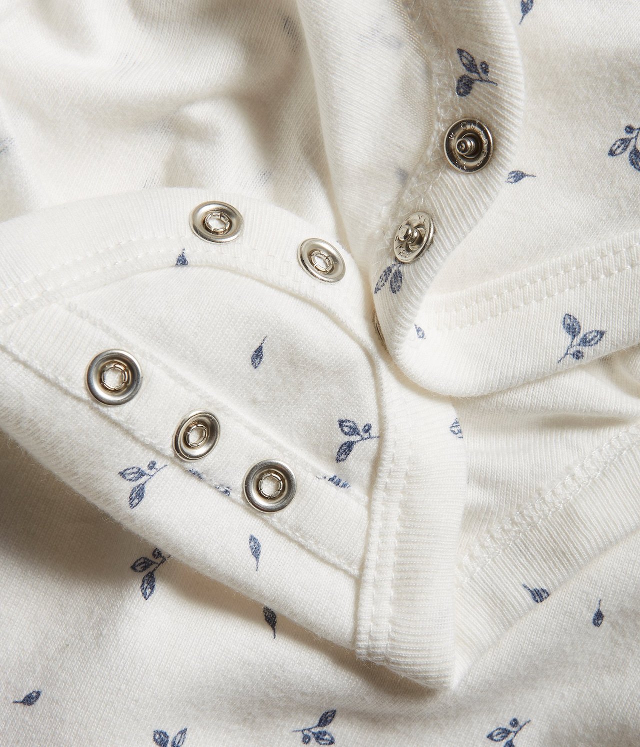 Nahaufnahme eines cremefarbenen Baumwoll-Baby-Bodys mit blauem Blattmuster und doppelten Druckknöpfen.
