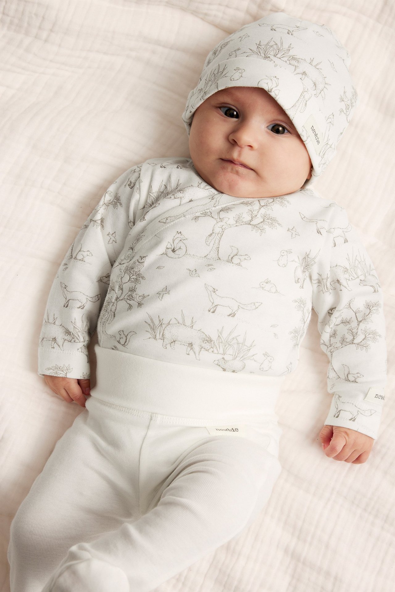 Baby in cremeweißem gemustertem Baumwoll-Wickelbody, passender Mütze und cremeweißer Hose.