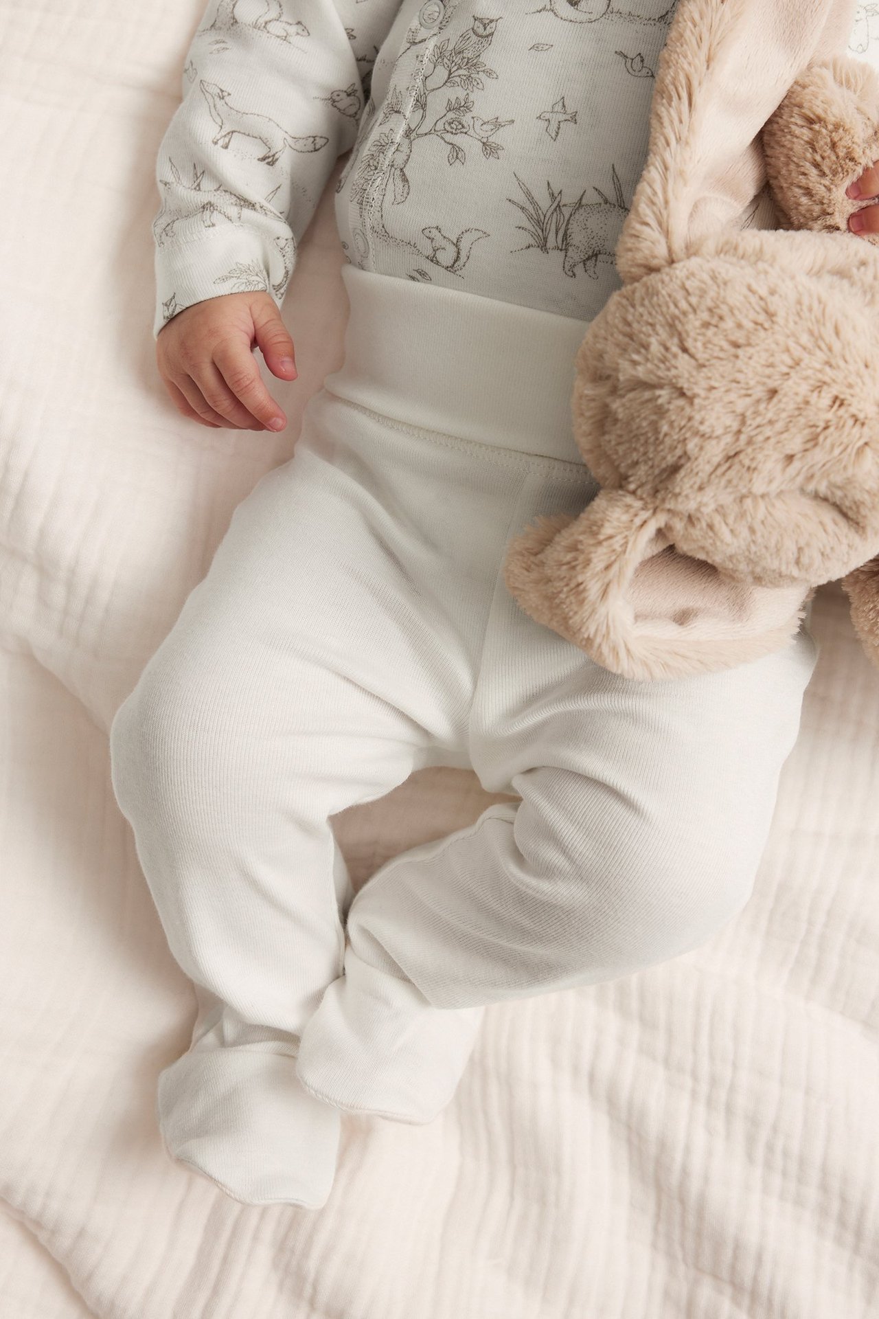 Ansicht von oben: Baby trägt off-white gemusterten Langarmbody, passende Hose mit Füßen, hält Plüschhasen.