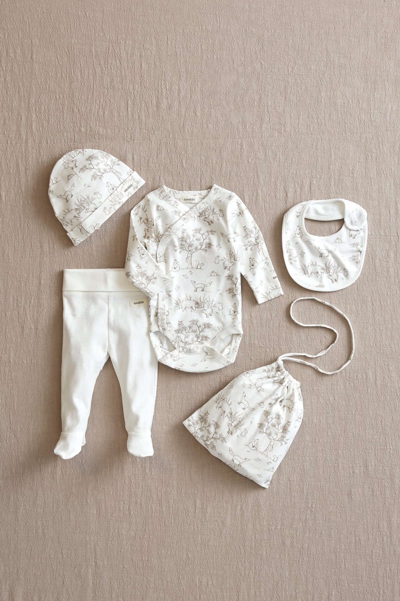 Von oben: Offwhite gemusterter Baby-Wickelbody, passender Hut, Lätzchen, Stoffbeutel und unifarbene Hosen mit Füßen.