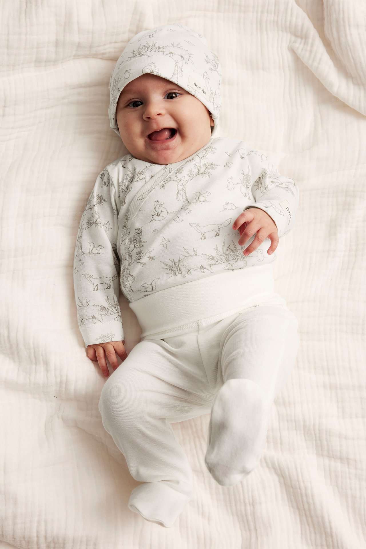 Vorderansicht eines lächelnden Babys in einem offwhite gemusterten Wickelbody, passender Mütze und unifarbenen Fußhosen.
