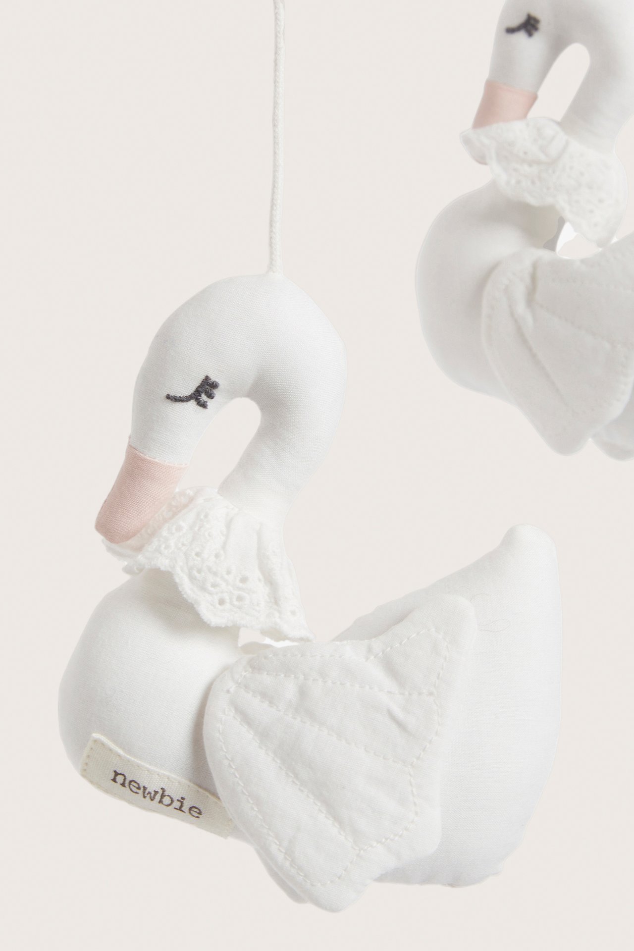 Close-up of an off-white cotton hanging mobile with two swan figures, featuring embroidered eyes and lace collars.