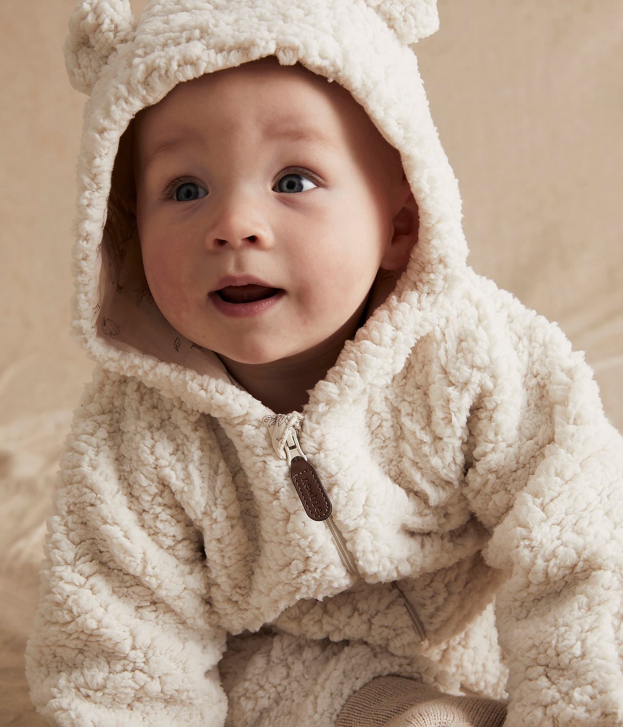 Close-up front view of a baby wearing a white pile overall with a hooded ear design and zipper.