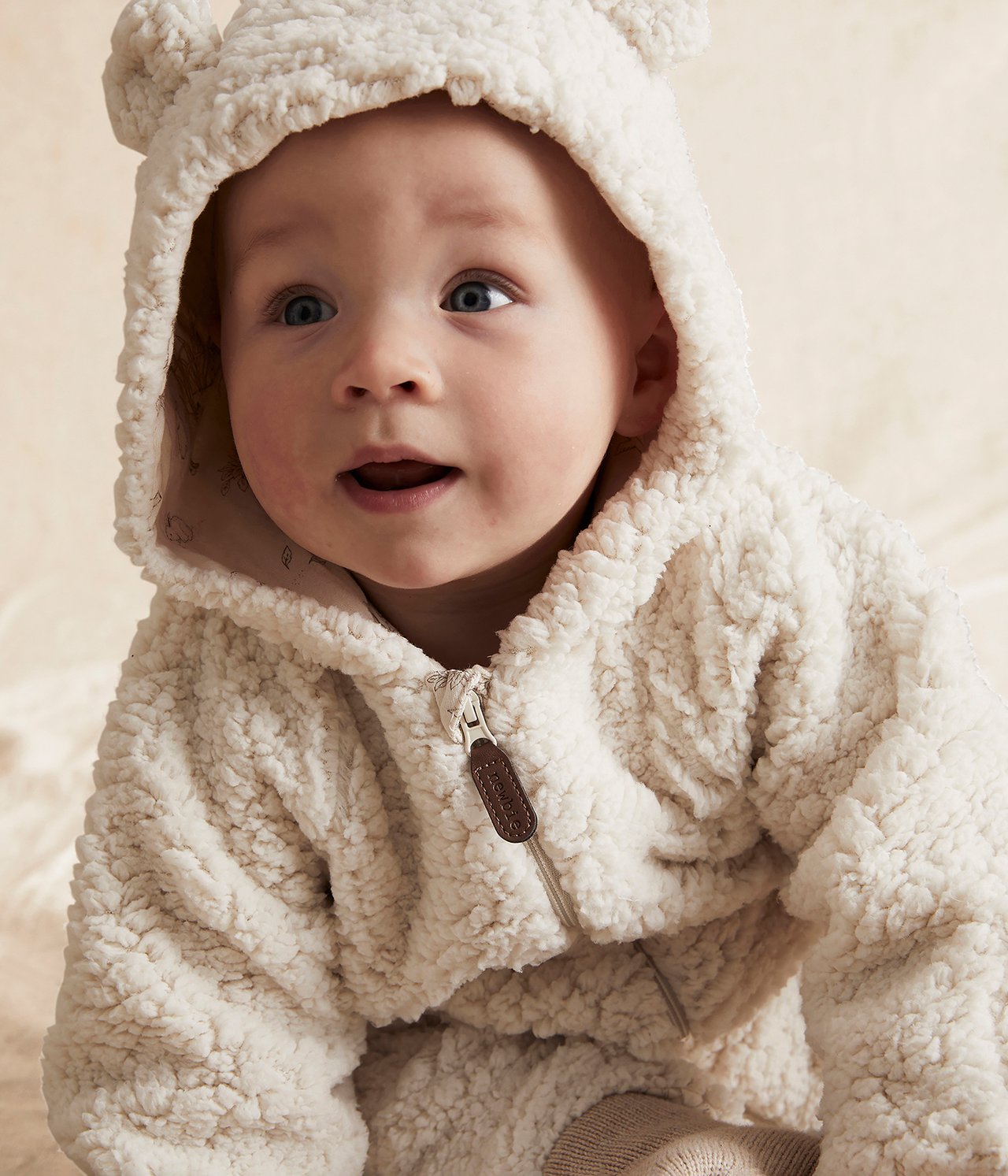 Nahaufnahme frontal eines Babys in einem cremefarbenen flauschigen Overall mit Reißverschluss und braunem Zipp.