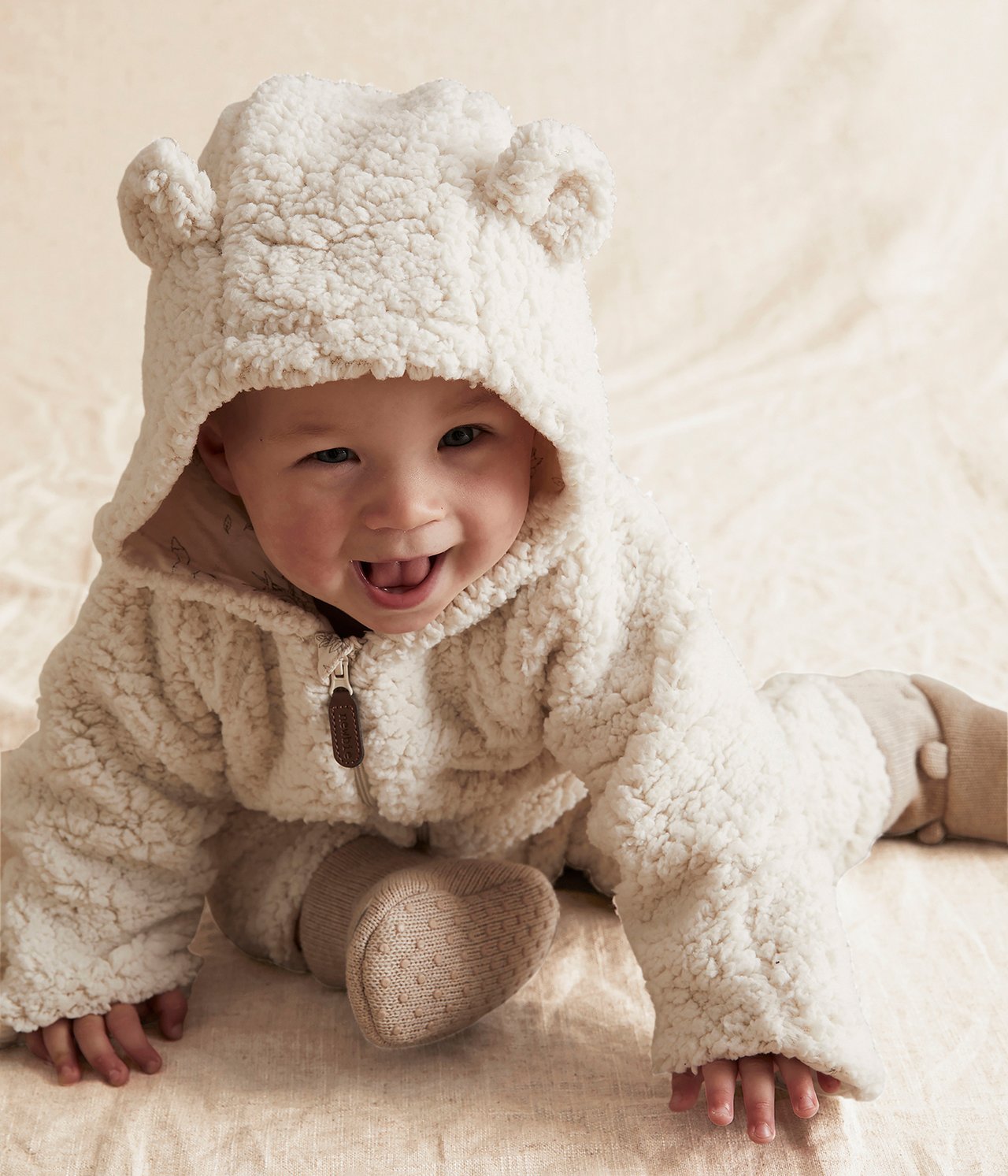 Front view of a baby in a cream fluffy hooded romper with bear ears and knitted booties.