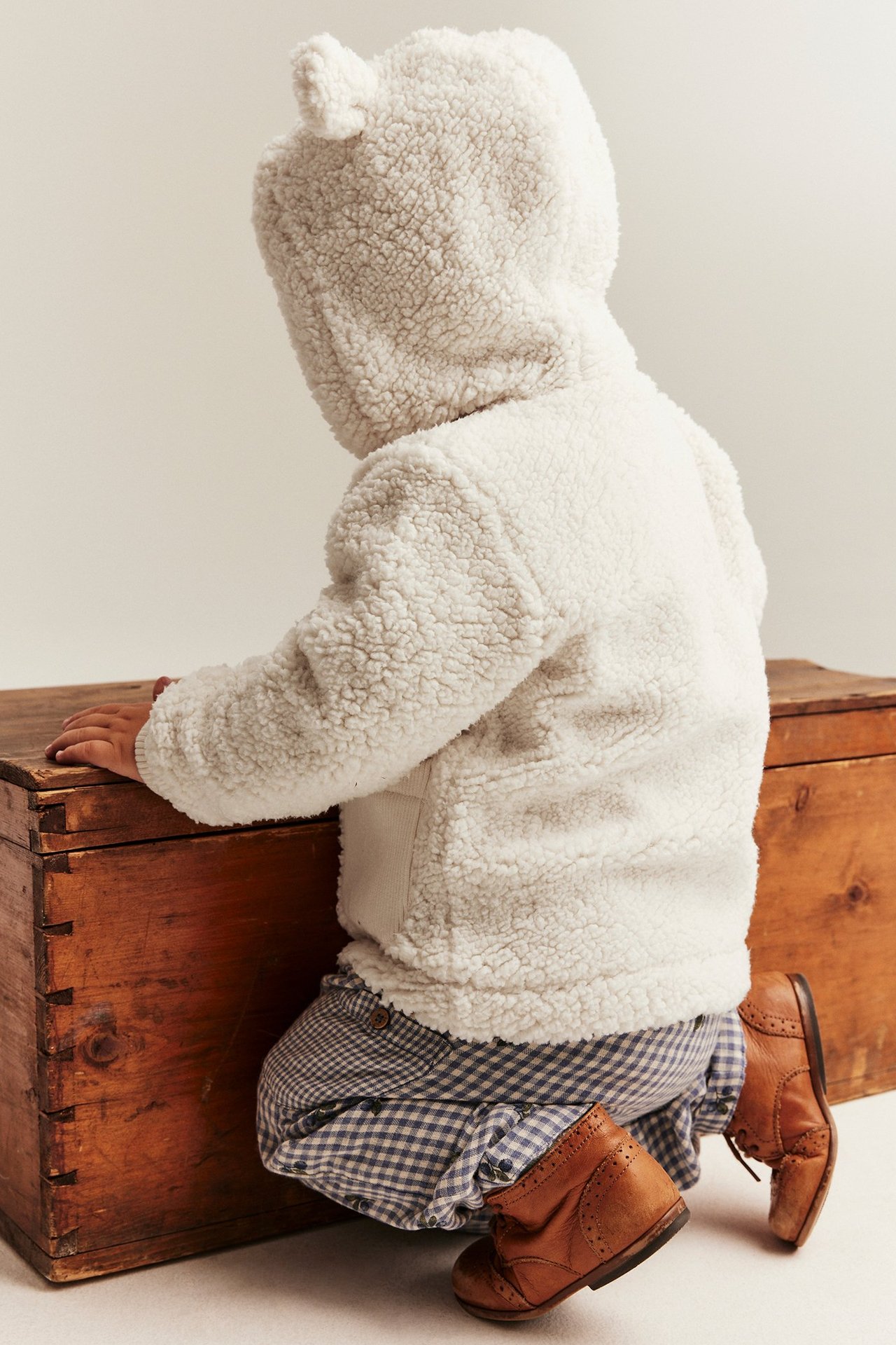 Rückenansicht Baby in weißer Teddy-Fleece-Kapuzenjacke mit Ohren, blauen Vichy-Shorts und braunen Lederstiefeln.