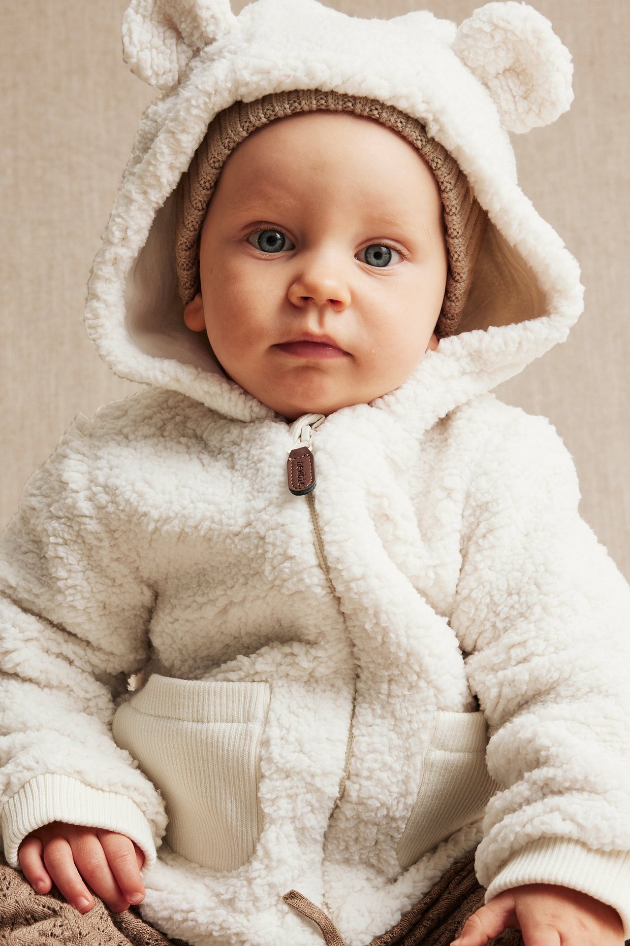 Close-up front view of a baby in a white teddy fleece jacket with bear ears, zipper, and light brown beanie.