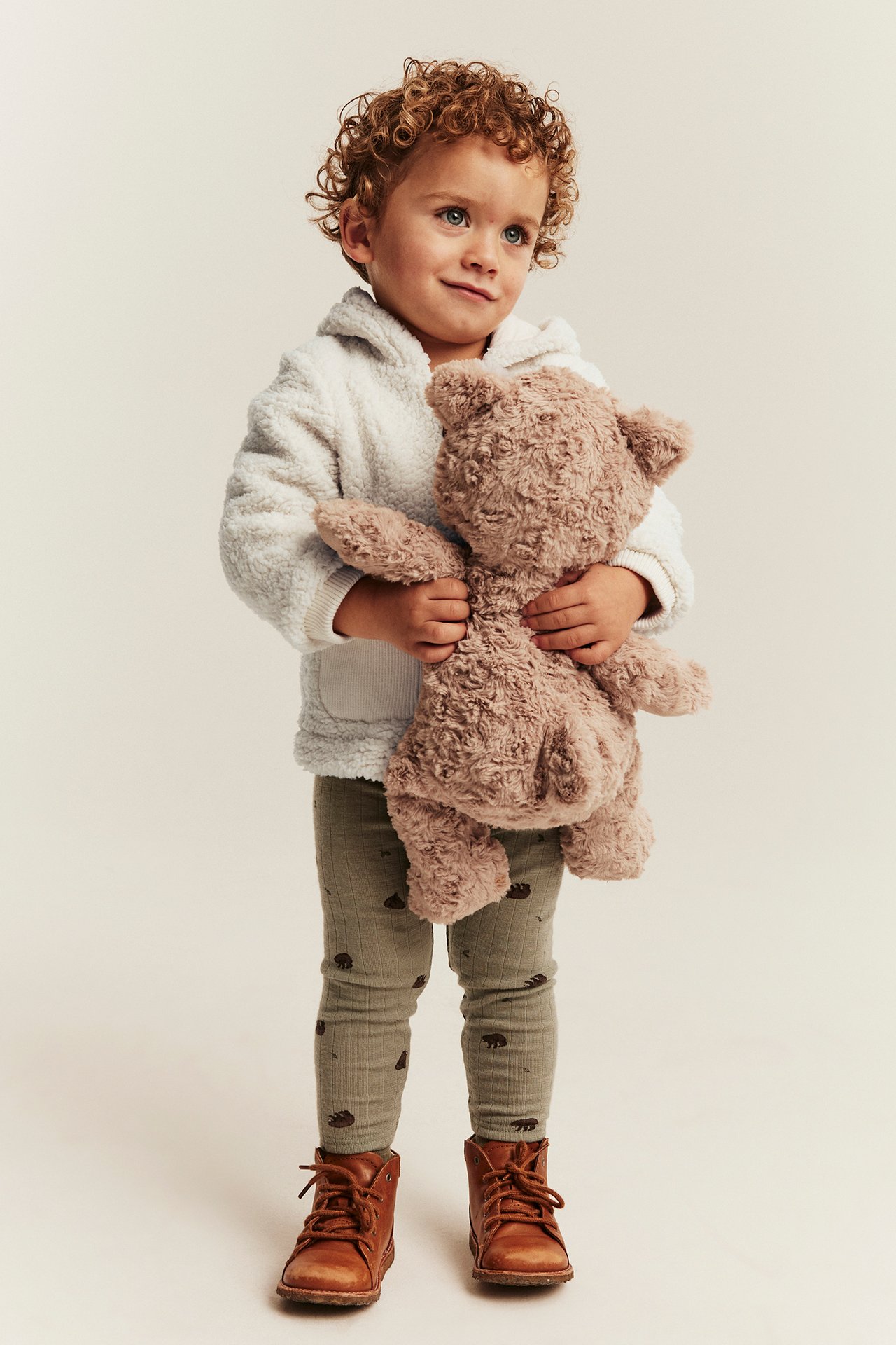 Front view: Kid in white fuzzy hoodie, olive green patterned leggings, brown boots, holding a brown teddy bear.