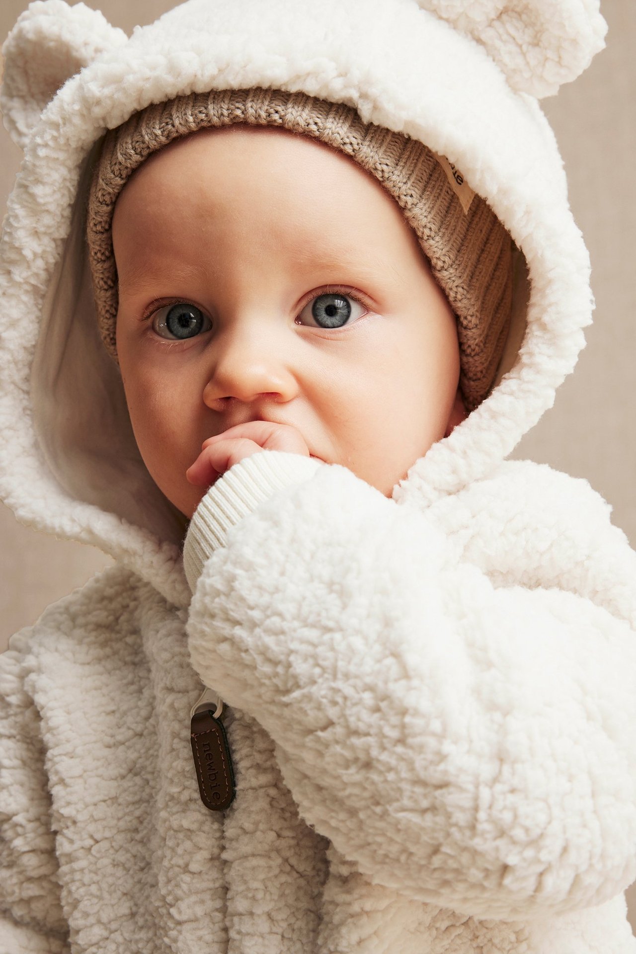 Close-up front view of a baby wearing a white teddy fur jacket with bear ears and a light brown beanie.