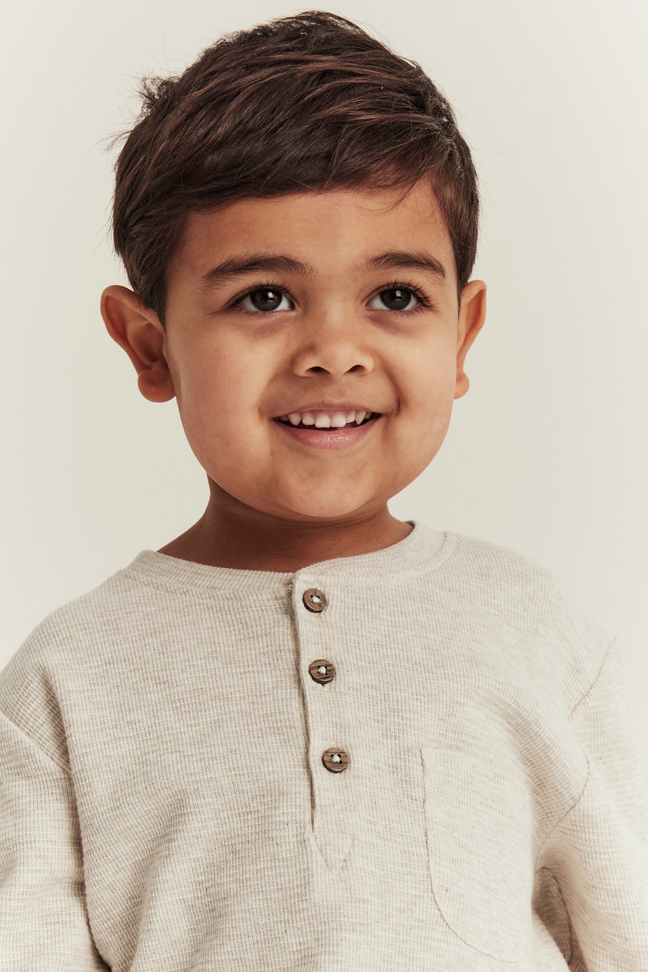 Vorderansicht eines lächelnden Kindes in einem beigemelange Waffelstrick-Langarmshirt mit Knöpfen und Brusttasche.