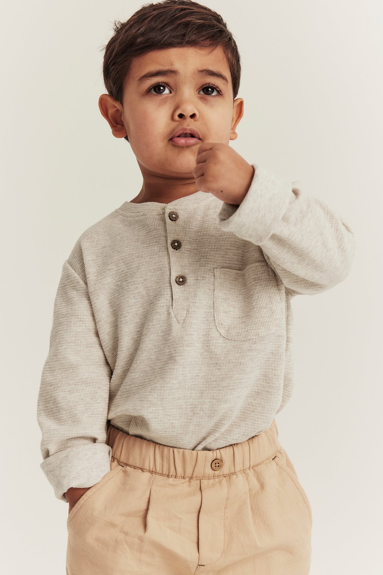 Vorderansicht: Kind in beigem Melange-Waffel-Grandad-Shirt mit Knöpfen, Tasche und hellbrauner Hose.