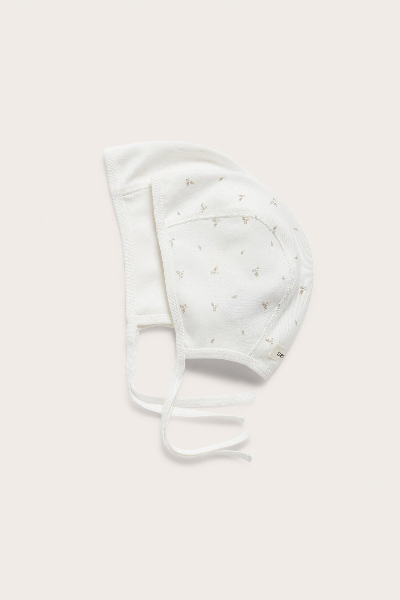 Flat lay of two light beige cotton baby bonnets: one patterned with leaves, one solid, both with ties.