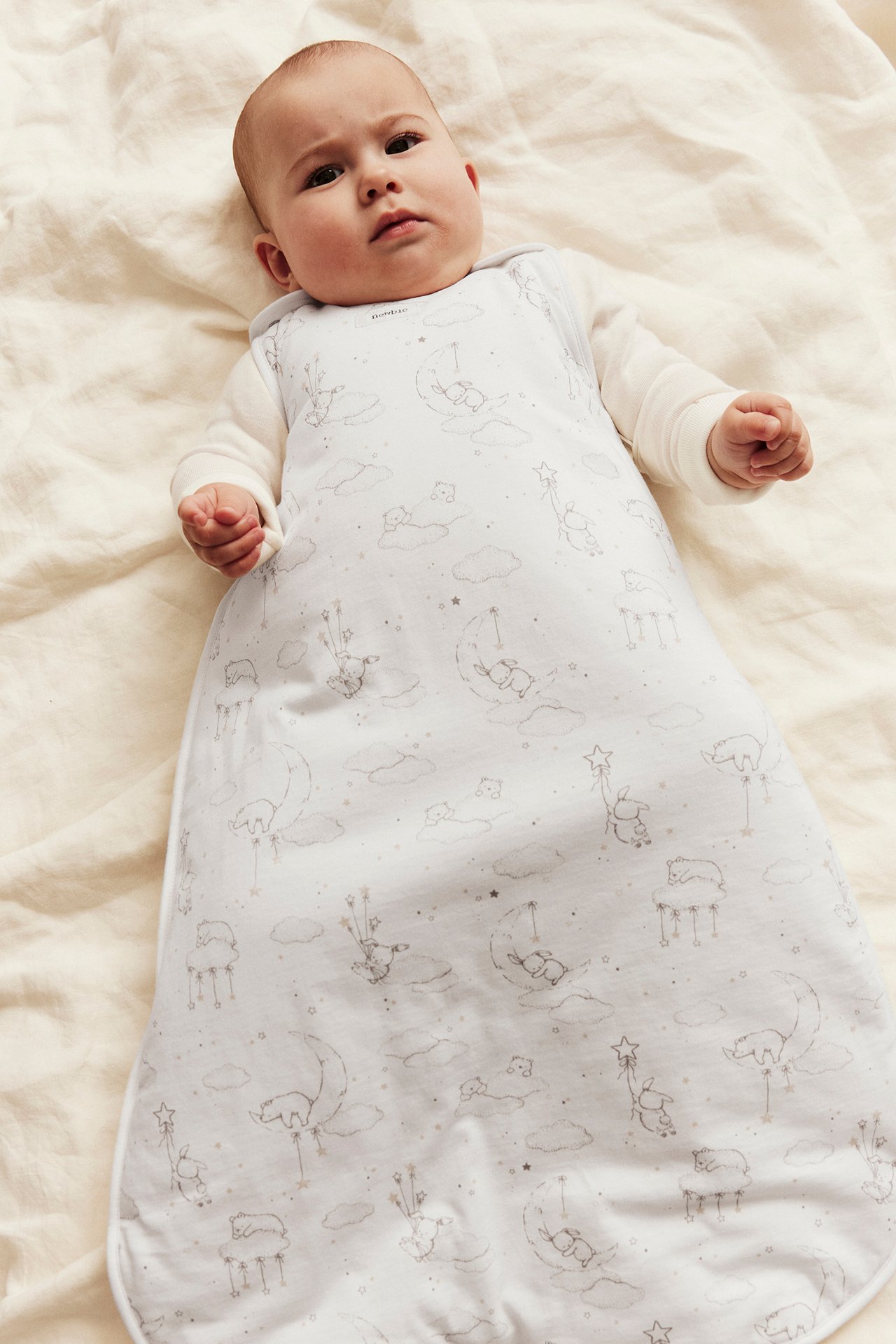 Top view: Baby in white sleep sack with animal print and stars, over a white long-sleeved top.