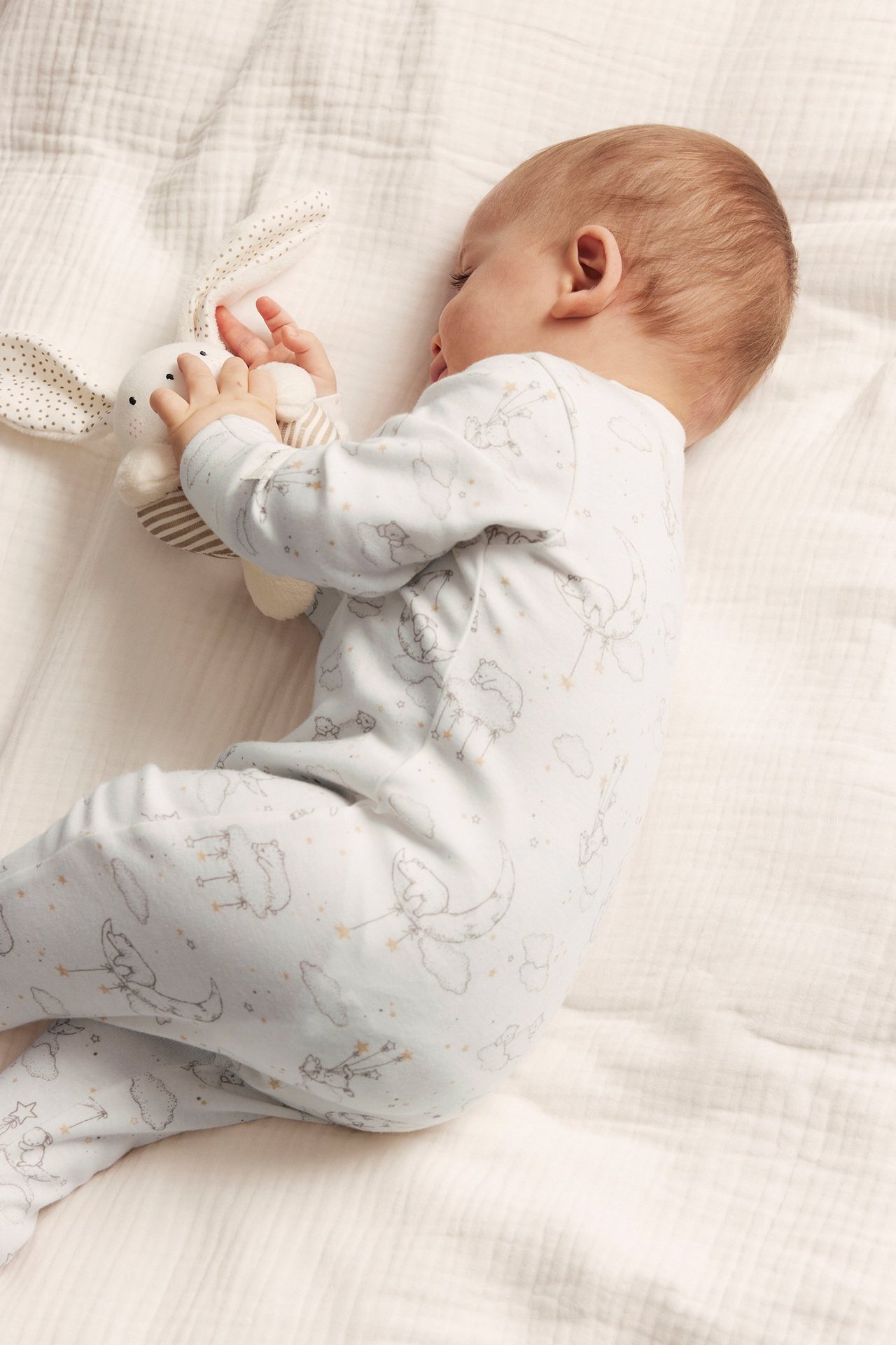 Side view of a baby in off-white cotton pajamas with bear and moon print, holding a white stuffed bunny.