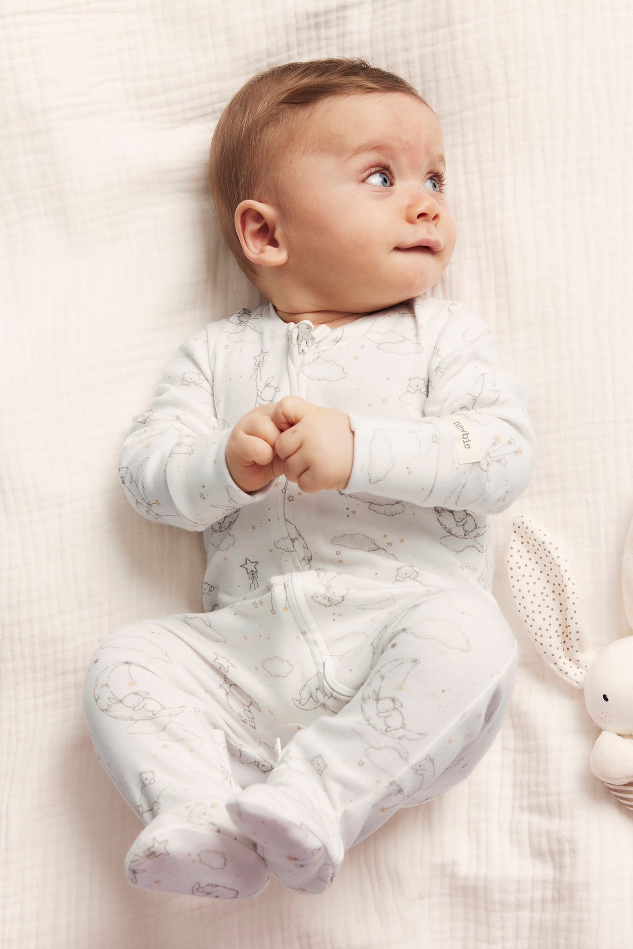 Top view of a baby in off-white cotton pyjamas with bear, moon, star pattern, zipper, footed. Bunny toy visible.