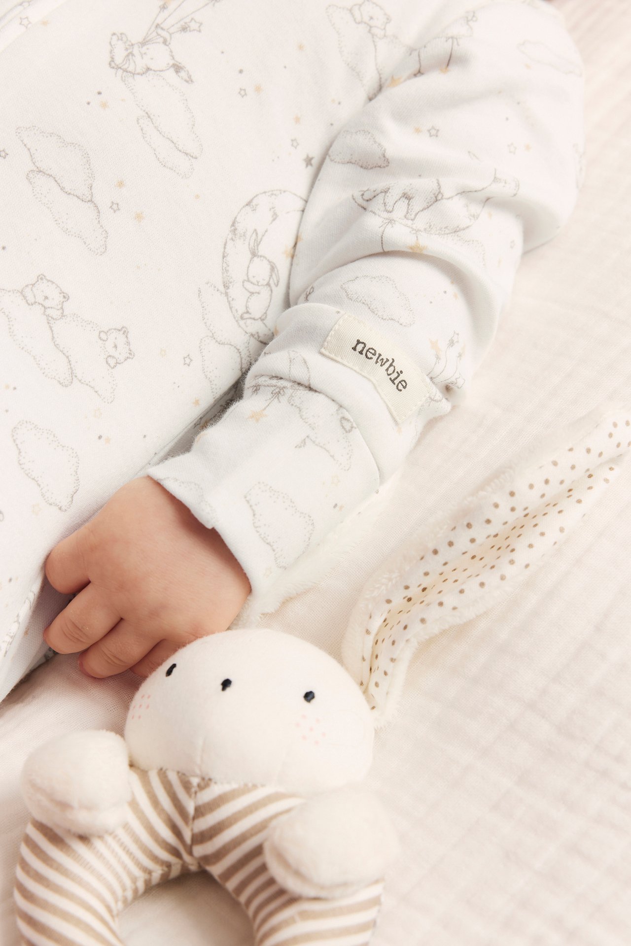 Close-up of baby's arm in offwhite cotton pajamas with animal pattern, next to a bunny rattle.